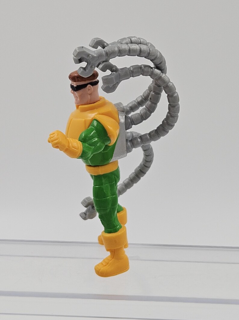 May include: A Dr. Octopus action figure. The figure is wearing a yellow and green outfit with grey mechanical arms extending from its back. The figure has brown hair and sunglasses.