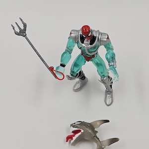 May include: A Spider-Man action figure in a teal and silver aquatic suit, holding a trident. A small, gray shark figure with a red tongue is also present. The figure has a red mask and silver boots.