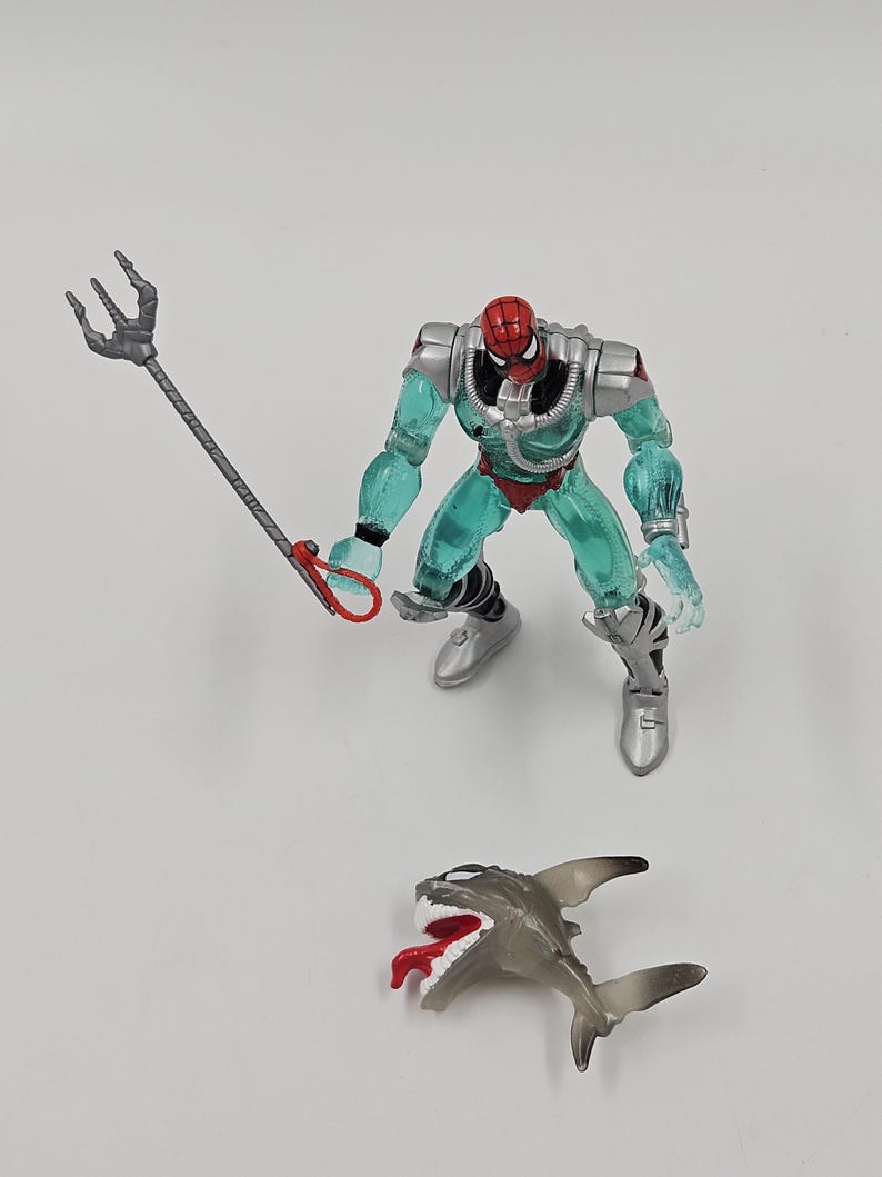 May include: A Spider-Man action figure in a teal and silver aquatic suit, holding a trident. A small, gray shark figure with a red tongue is also present. The figure has a red mask and silver boots.