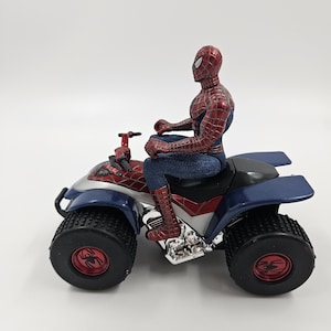 May include: A Spider-Man action figure sits atop a blue, red, and silver ATV quad bike. The ATV has large black tires with red spider emblems. The Spider-Man figure is in a classic red and blue suit.