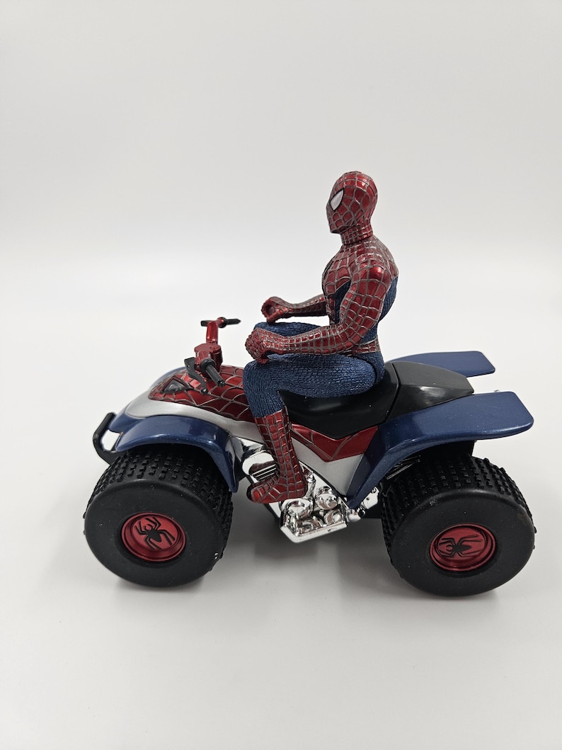May include: A Spider-Man action figure sits atop a blue, red, and silver ATV quad bike. The ATV has large black tires with red spider emblems. The Spider-Man figure is in a classic red and blue suit.