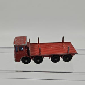 May include: A vintage red toy truck with a flatbed trailer. The truck has a detailed cab with windows and four black wheels. The trailer has vertical supports and is also red. A classic toy vehicle.