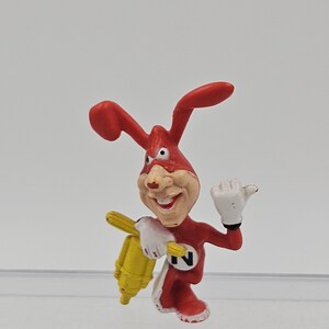 May include: A small red cartoon character figurine with long ears, a tan face, and a wide grin. The figure wears a red suit with a white circle containing a black letter N on the chest, white gloves, and holds a yellow object in one hand.