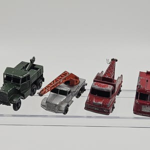 May include: Four vintage toy trucks in a row. The trucks are in various colors, including green, silver, and red. One truck has a crane, and another has a lifting arm. The trucks are on a clear surface.