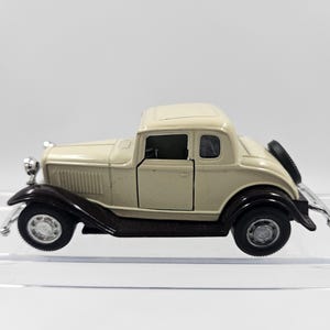 Könnte beinhalten: Ein Modellauto eines klassischen Oldtimers. Das Auto ist cremefarben mit schwarzen Kotflügeln und Rädern. Ein Reserverad ist hinten montiert. Das Auto hat Chromakzente an den vorderen und hinteren Stoßfängern.