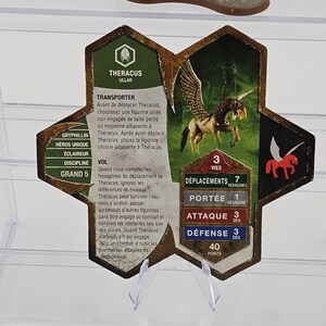 Może przedstawiać: Sześciokątny element gry z ilustracją skrzydlatego konia, o nazwie "Theracus". Element ma zielone, brązowe i białe elementy, z tekstem w języku angielskim i francuskim. Jest prezentowany na przezroczystym stojaku.