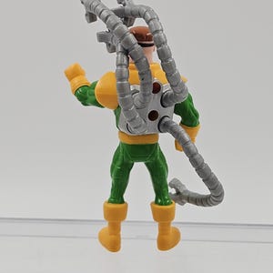 May include: A plastic Doctor Octopus action figure. The figure is dressed in a green and yellow suit, with a brown head and four articulated grey mechanical arms. The figure is standing on a clear surface. The toy is a comic book villain.