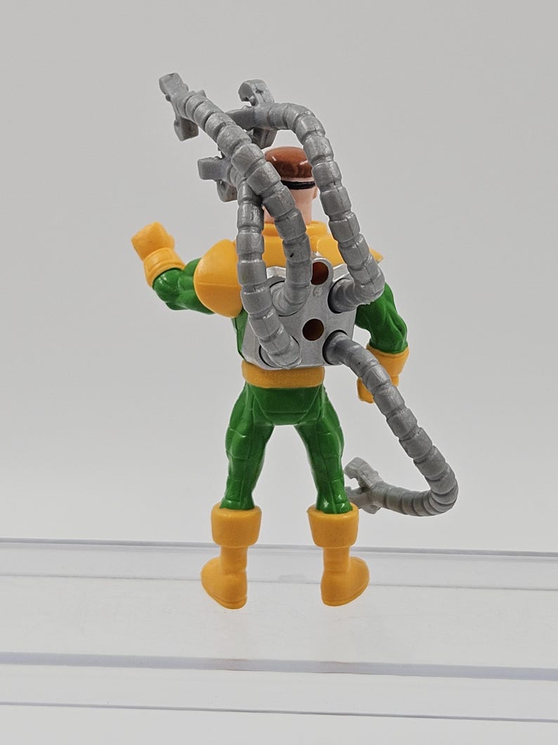 May include: A plastic Doctor Octopus action figure. The figure is dressed in a green and yellow suit, with a brown head and four articulated grey mechanical arms. The figure is standing on a clear surface. The toy is a comic book villain.