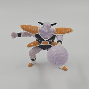 May include: A purple and white action figure of a character from the Dragon Ball Z anime series. The figure is wearing a black and orange suit with a purple and white energy ball in its hand.