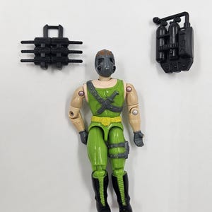 May include: An action figure in green and tan with a black mask and accessories. The figure has a green top and trousers, with black boots. Accessories include a black backpack and a black weapon. The figure is articulated and has a vintage style.