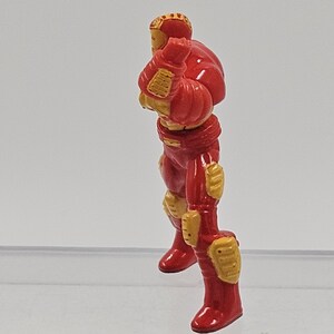 May include: A small, molded figurine of a character in a red and gold armored suit. The figure is depicted in a standing, side-profile pose with arms bent at the elbows and hands positioned near the chest area, featuring detailed plating.