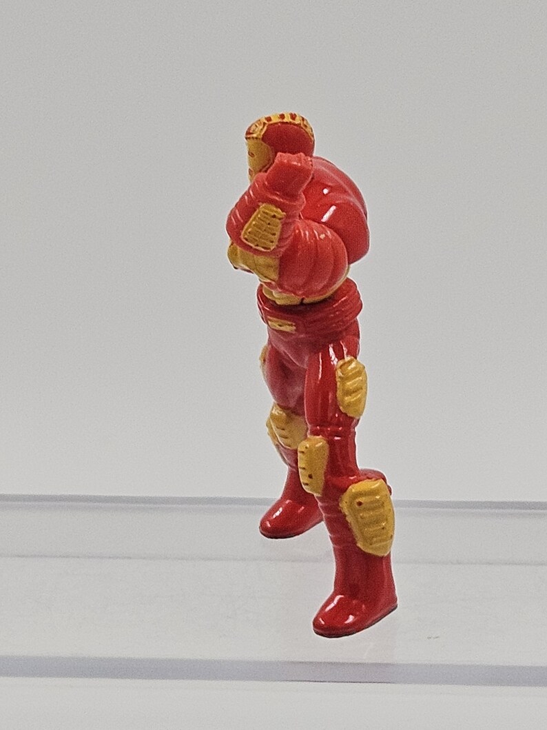 May include: A small, molded figurine of a character in a red and gold armored suit. The figure is depicted in a standing, side-profile pose with arms bent at the elbows and hands positioned near the chest area, featuring detailed plating.