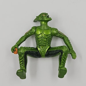 Op de afbeelding: Groene actiefiguur van een fictief personage, in zittende positie. De figuur heeft een gedetailleerd, gestructureerd ontwerp met zwarte accenten en houdt een klein oranje object vast. Het gezicht van de figuur heeft een dreigende uitdrukking.