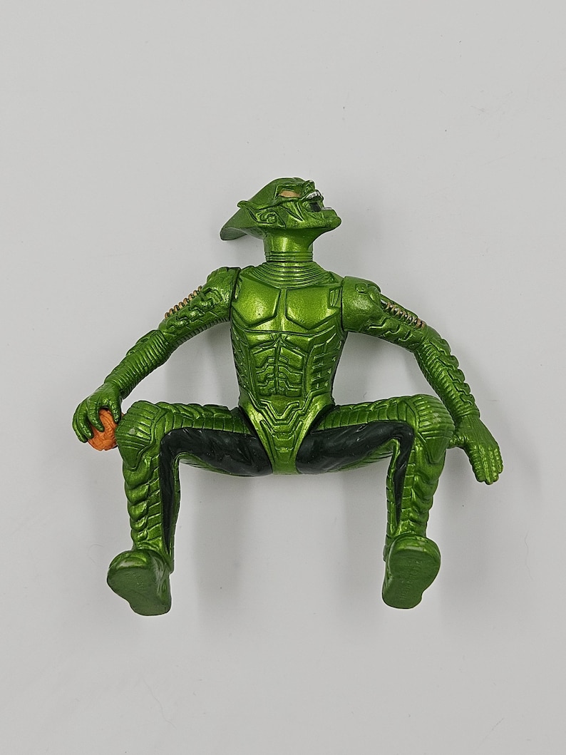 Op de afbeelding: Groene actiefiguur van een fictief personage, in zittende positie. De figuur heeft een gedetailleerd, gestructureerd ontwerp met zwarte accenten en houdt een klein oranje object vast. Het gezicht van de figuur heeft een dreigende uitdrukking.