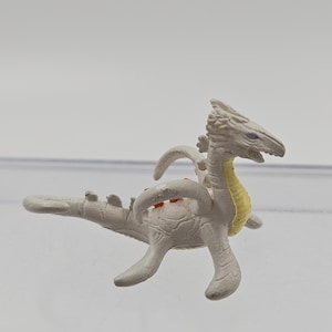 Peut inclure: Petite figurine de dinosaure grise avec un long cou et une longue queue, et quatre petites pattes. Le dinosaure a un ventre jaune et des taches rouges sur le dos. La figurine est en plastique et est probablement un jouet.