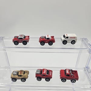 Puede incluir: Una colección de coches de juguete en miniatura, incluyendo una camioneta roja, un coche deportivo dorado y una furgoneta blanca, todos con el logotipo de Coca-Cola. Los coches se exhiben en una estantería transparente de varios niveles. Los coches miden unos 5 cm de largo.