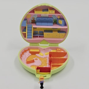 Può includere: Un giocattolo vintage a forma di cuore, aperto per rivelare stanze in miniatura con mobili. La casa delle bambole è verde chiaro e rosa, con una piccola bambola davanti. L'interno presenta un divano blu, una cucina gialla e scaffali arancioni.