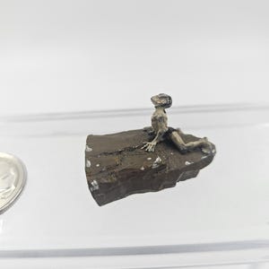 May include: A small, detailed figurine of a person reclining on a rock. The figure is a light brown color, with dark accents. The rock is a dark brown with white speckles. The figurine is set against a white background.