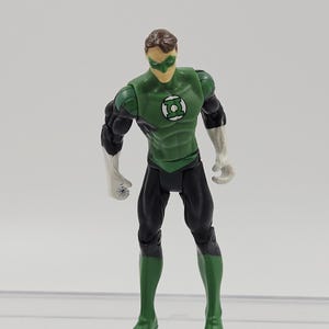May include: A Green Lantern action figure. The figure is wearing a green and black costume with a white emblem on the chest. The figure has a green mask and brown hair. The figure is standing with its arms at its sides.