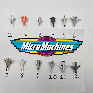 Puede incluir: Una colección de doce aviones de juguete en miniatura Micro Machines, cada uno con diseños y colores únicos, dispuestos sobre una superficie blanca. El logotipo de Micro Machines es visible. Los números del 1 al 12 están escritos debajo.