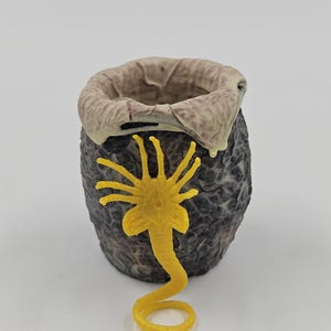 Puede incluir: Un huevo detallado y texturizado, de color gris oscuro, con un borde marrón claro y un facehugger amarillo. El huevo tiene una superficie rugosa y con bultos, y el facehugger está enrollado.