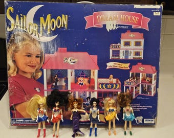 May include: Sailor Moon Dream House furniture set with dolls. The box features a girl, a crescent moon, and a pink and blue house. The set includes several Sailor Moon character dolls.