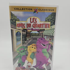 May include: A VHS tape featuring the characters Barney, Baby Bop, and BJ. The cover art shows the characters crossing a street in front of a town scene. The title "Les Amis du Quartier" is displayed prominently.