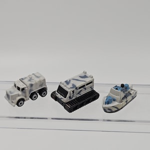 May include: Three miniature toy vehicles featuring a white and blue camouflage pattern. The set includes a six-wheeled truck, a tracked vehicle with a boxy cabin, and a small boat with blue accents, all displayed together as a collection of models.