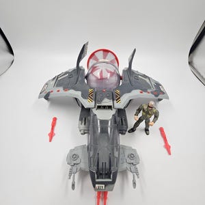 May include: A gray and red toy spaceship with a clear cockpit and red missile accessories. A small action figure in a flight suit stands nearby. The spaceship has a red and clear dome on top.