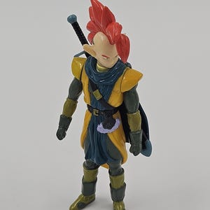 May include: A red-haired action figure with a sword strapped to its back. The figure is wearing a yellow and green outfit with a blue cape.