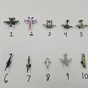 Puede incluir: Una colección de diez aviones de juguete en miniatura, cada uno con diseños y esquemas de color únicos. Los aviones están numerados del 1 al 10 y están dispuestos sobre una superficie blanca. Los aviones varían en tamaño y estilo, algunos con diseños de hélice.