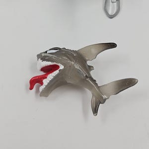 May include: A translucent gray shark toy with a red tongue and white teeth. The toy shark has a detailed design, including fins and a tail. The image also shows parts of a silver action figure.