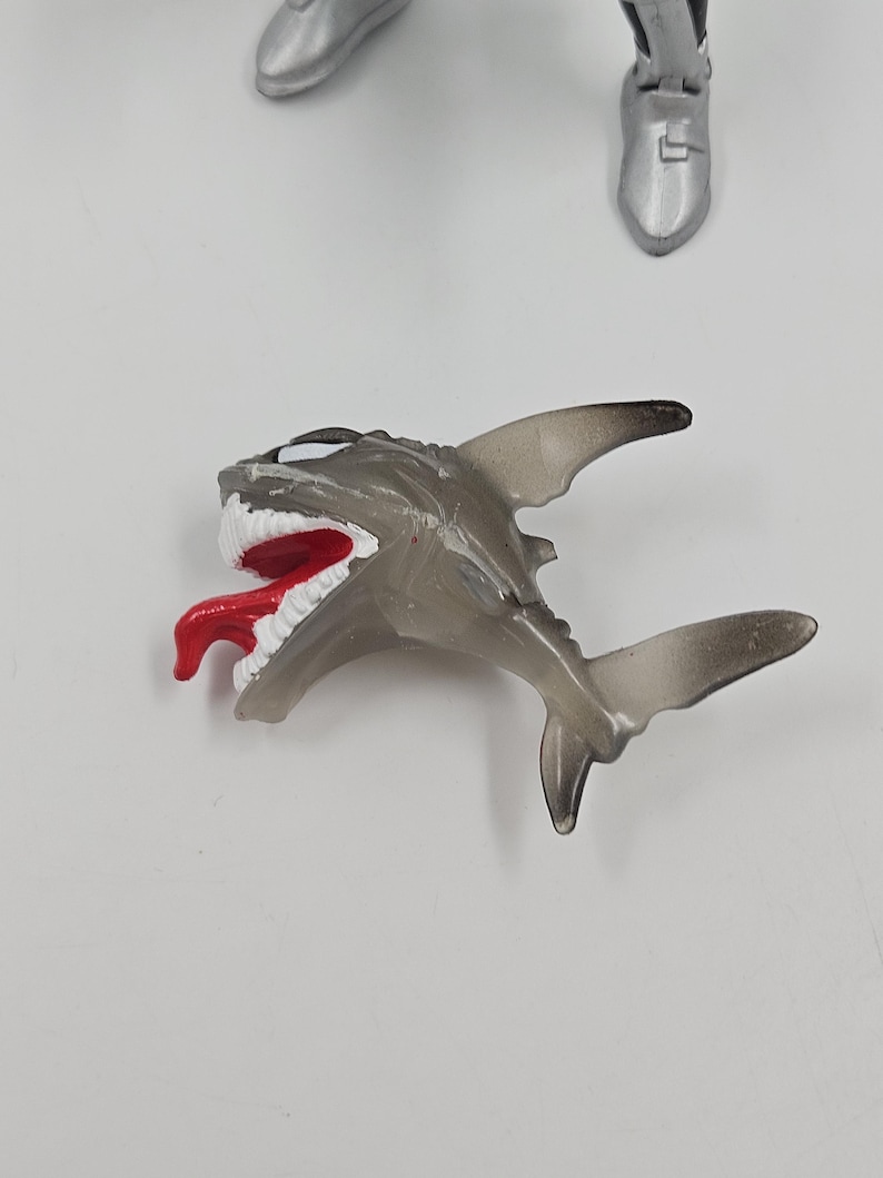 May include: A translucent gray shark toy with a red tongue and white teeth. The toy shark has a detailed design, including fins and a tail. The image also shows parts of a silver action figure.