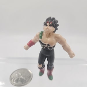 May include: A small plastic action figure of a muscular character with black hair and a determined expression. The figure wears a black and green outfit with red accents. A silver coin is next to the figure.