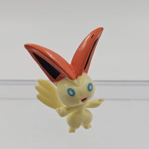 Peut inclure: Une petite figurine jaune avec des accents orange et noirs. La figurine a de grandes oreilles orange, des yeux bleus et une petite queue pointue. Elle est posée avec un bras tendu.