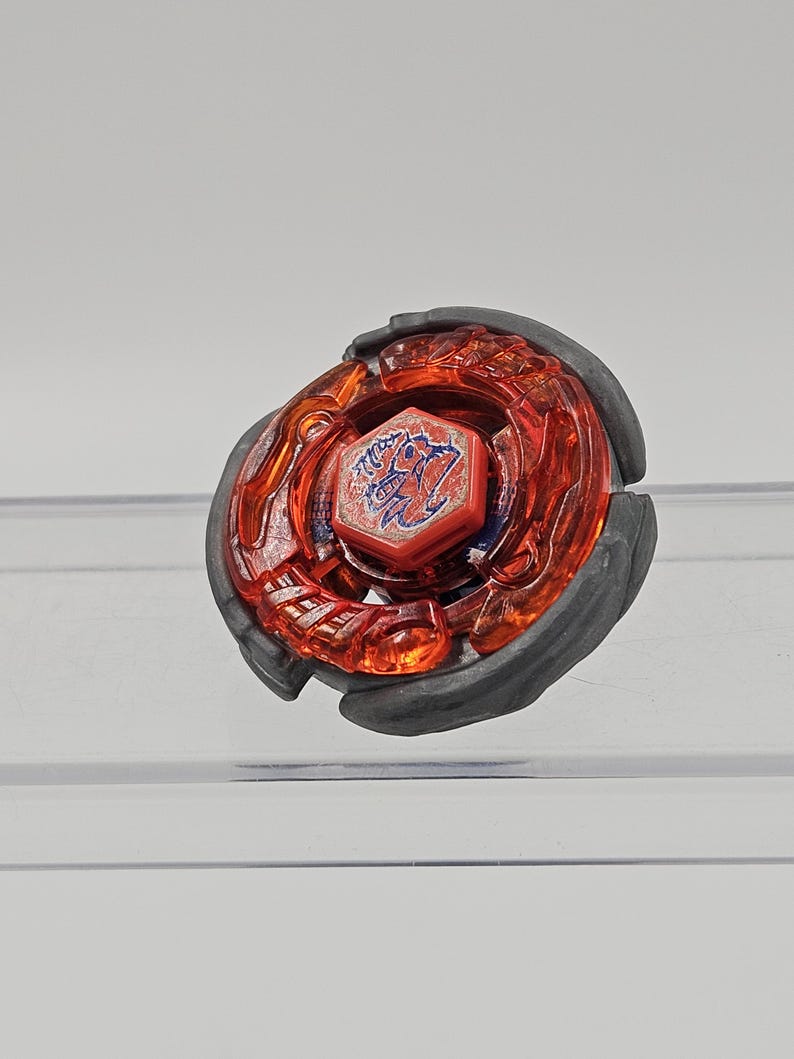 Peut inclure: Un toupie rouge et noir avec un embl&egrave;me bleu central. Le dessus a une couche ext&eacute;rieure rouge transparente et un anneau ext&eacute;rieur gris fonc&eacute;. L'embl&egrave;me pr&eacute;sente un design stylis&eacute;.