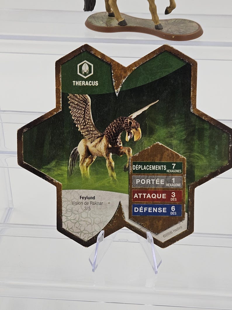 Może przedstawiać: Karta do gry z ilustracją skrzydlatego konia, o nazwie "THERACUS". Karta zawiera statystyki gry i tekst "Feylund Vision de Raknar 3/5". Tło jest zielono-brązowe, z informacją o prawach autorskich Hasbro.