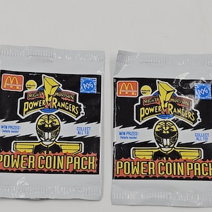 Könnte beinhalten: Zwei ungeöffnete Power Rangers Power Coin Pack-Päckchen. Jedes Päckchen hat einen schwarzen Hintergrund mit gelbem und rotem Text. Die Päckchen zeigen das Power Rangers-Logo, eine Power Ranger-Helm-Grafik und den Text "POWER COIN PACK".