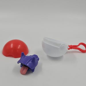 Puede incluir: Un juguete desmontado que presenta una cúpula roja, un recipiente blanco con un clip rojo y un personaje morado con una lengua rosa. El juguete tiene un diseño de dibujos animados.