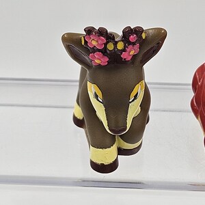May include: A small brown deer figurine with yellow accents and a pink flower crown. The deer is sitting on its hind legs with its front legs extended.