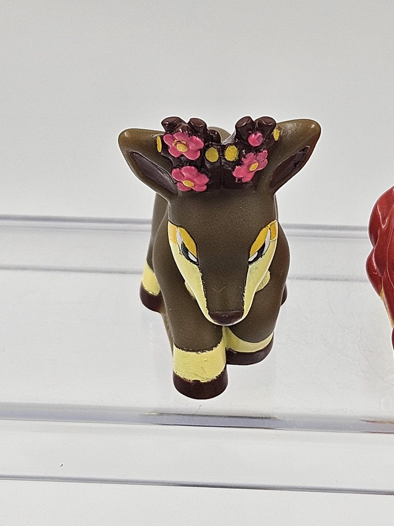 May include: A small brown deer figurine with yellow accents and a pink flower crown. The deer is sitting on its hind legs with its front legs extended.