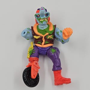 May include: A vintage action figure of a mutated humanoid with blue skin, a green face, and orange hair. The figure wears a brown vest, purple pants, and red boots. It has a wheel attached to one leg and an orange fist.