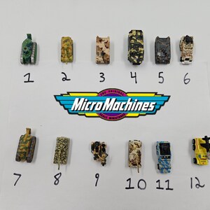 Peut inclure: Une collection de douze véhicules miniatures Micro Machines, comprenant des chars, des camions et des hélicoptères, dans divers motifs et couleurs de camouflage. Le logo Micro Machines est visible.