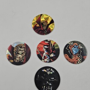 May include: Five circular collectible tokens featuring comic book characters. The tokens display colourful illustrations of characters with detailed facial features and costumes. The tokens are arranged against a plain background.