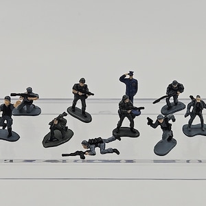 May include: A collection of miniature toy soldiers in various action poses. The figures are primarily black and gray, with some wearing helmets and carrying weapons. One figure is in a blue uniform. They are arranged on a clear, horizontal surface.