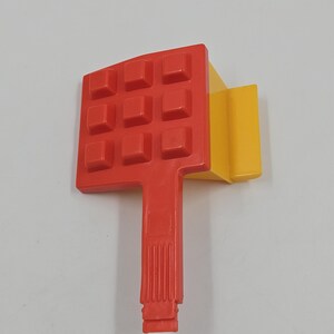 Puede incluir: Un juguete de plástico rojo y amarillo. La parte roja tiene una cuadrícula de protuberancias cuadradas, un asa y una base rectangular. La parte amarilla es un bloque rectangular unido a la pieza roja. El juguete está sobre un fondo blanco.