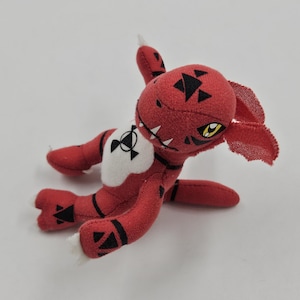 Peut inclure: Une peluche rouge représentant une créature avec des marques noires et des accents blancs. Le jouet a des yeux jaunes, des dents blanches et un emblème blanc et noir sur la poitrine. La créature est allongée sur une surface blanche.