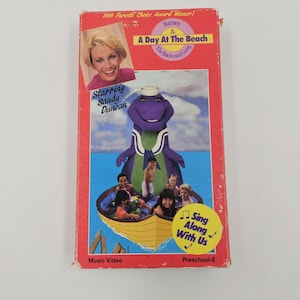 May include: A red VHS tape featuring Barney the dinosaur and the Backyard Gang. The cover includes the title "A Day At The Beach", the text "Starring Sandy Duncan", and the phrase "Sing Along With Us".