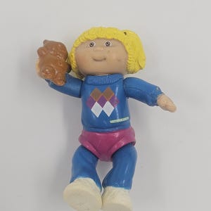 May include: A vintage Cabbage Patch Kids doll figurine. The doll has yellow hair, a blue sweater with a diamond pattern, pink shorts, and blue pants. It is holding a brown teddy bear. The doll's face is detailed with painted features.