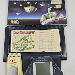 May include: A vintage Golf Challenge electronic game set. The box features a golf course illustration. The set includes a handheld game console, a course guide, and a golf ball. The game console has a screen and buttons.